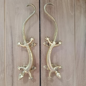 Peut inclure: Deux poignées de porte en laiton en forme de lézards stylisés. Les poignées sont incurvées avec un dos texturé et des détails décoratifs. Les pattes et la queue du lézard sont visibles, montées sur une surface en bois clair.