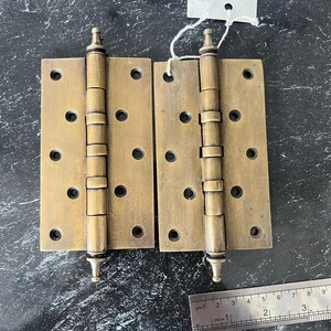 May include: Two antique brass door hinges. Each hinge has a rectangular plate with four evenly spaced holes and a cylindrical hinge pin. The hinges are a warm, aged brass color, suggesting vintage hardware. A ruler is in the bottom right corner.