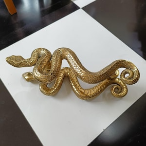 Peut inclure: Une poignée de porte dorée en forme de serpent. La poignée présente des écailles détaillées et une queue enroulée. La tête du serpent est à une extrémité, et le corps se courbe gracieusement sur la surface.