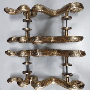 May include: Four brass-coloured door handles, each shaped like a snake. The snakes curve and coil, with detailed scales and heads. The handles are mounted on a metal rod with circular accents, creating a unique and decorative design.