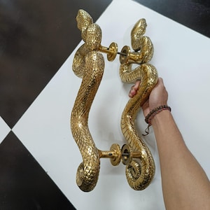 Könnte beinhalten: Zwei Türgriffe aus Messing in Form von Schlangen. Die goldfarbenen Griffe weisen detaillierte Schuppen und Schlangenkopf-Designs auf. Sie sind auf einer weißen Oberfläche montiert. Die Griffe sind etwa 30 cm lang.