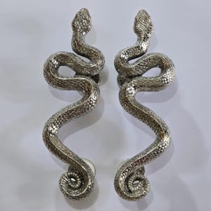 Peut inclure: Deux poignées de porte argentées en forme de serpents. Les poignées présentent des écailles texturées et des queues enroulées. Les têtes de serpent sont relevées et les poignées sont montées sur des bases rondes argentées.