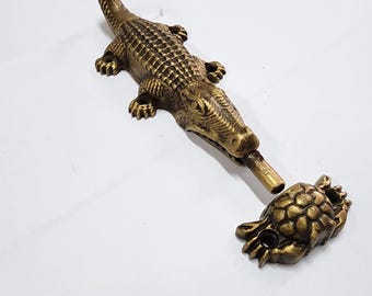 May include: A gold-toned metal doorstop shaped like a crocodile with a detailed textured body. The crocodile's mouth is open and a separate gold-toned metal turtle is attached to the crocodile's mouth.