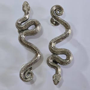 Peut inclure: Deux poignées de porte argentées en forme de serpents. Les poignées présentent des détails d'écailles texturées et des corps enroulés. Les têtes de serpent sont en haut et en bas, fixées sur des bases rondes argentées. Longueur approximative de 30 cm.