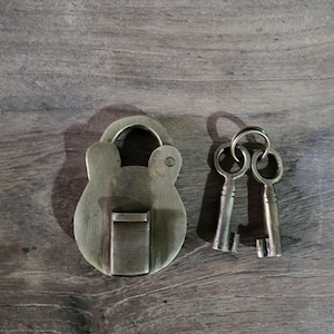 May include: Antique brass padlock with two matching keys. The padlock has a rounded shape with a keyhole on the side and a small latch on the front. The keys are small and have a simple design.