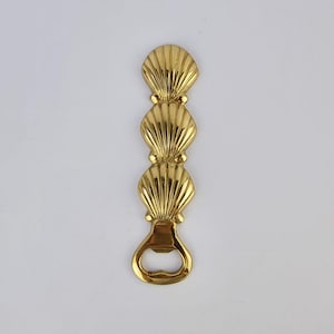 May include: A gold-coloured bottle opener with a design of three stacked seashells. The opener has a loop at the bottom for leverage and a decorative, coastal-themed aesthetic. The seashells are detailed with ribbed textures.