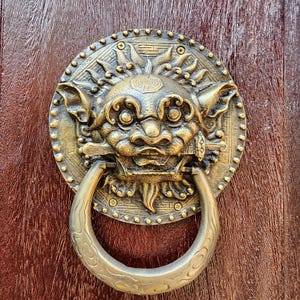 May include: A gold-toned door knocker with a detailed design of a mythical creature with a ring handle. The knocker is mounted on a wooden door.