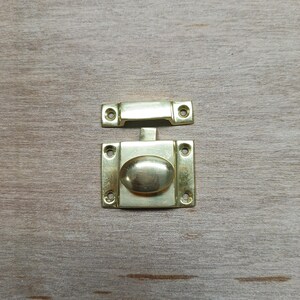 May include: A gold-toned metal latch with a rectangular base and a rounded handle. The latch is designed to be used on a door or cabinet.