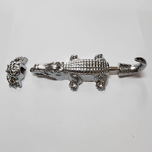 May include: A silver-toned alligator-shaped door latch with a hook on the tail. The latch is in three pieces, with the alligator body, a separate tail hook, and a separate mounting plate.