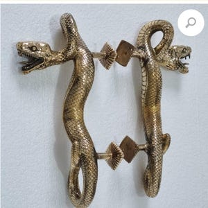 Może przedstawiać: Para złotych klamki do drzwi w kształcie węży z detalicznymi łuskami. Klamki są przymocowane do kwadratowej metalowej płytki z ozdobnym wzorem.