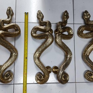 Pode incluir: Três pares de puxadores de porta dourados, cada um com um design detalhado de cobra. As cobras estão enroladas e entrelaçadas, com suas cabeças formando a parte superior da maçaneta. Uma fita métrica amarela é colocada verticalmente entre as maçanetas.