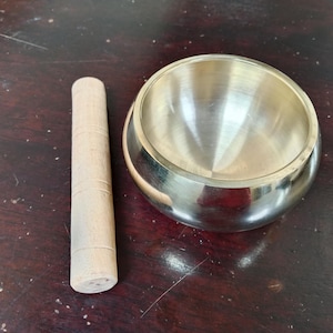 May include: A silver singing bowl with a wooden striker. The bowl is round and has a smooth, reflective surface. The striker is cylindrical and has a textured surface.