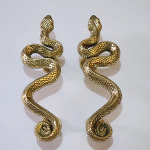 Puede incluir: Un par de tiradores de puerta de serpiente de color dorado. Los tiradores están hechos de metal y tienen un diseño de serpiente realista. Las serpientes están enroscadas en forma de S y tienen escamas detalladas.