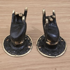 Könnte beinhalten: Zwei antike Schrankgriffe in Form von Händen, die eine Rock-and-Roll-Geste ausführen. Die Griffe sind dunkelbronze mit goldenen Akzenten an den Fingerspitzen und am Sockel. Jeder Griff ist auf einer dekorativen kreisförmigen Basis montiert.