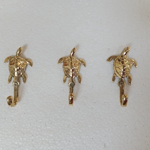 Könnte beinhalten: Drei goldfarbene Metall-Schildkrötenhaken. Jeder Haken hat oben ein Schildkröten-Design mit einem gebogenen Haken darunter. Die Haken sind zum Aufhängen von Gegenständen gedacht und verleihen eine dekorative Note.