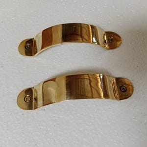 May include: Two polished brass cabinet handles with rounded ends and a slight curve. Each handle has a single screw visible on the side.