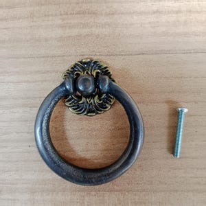 Puede incluir: Una manija de cajón de anillo negro con un diseño floral dorado decorativo y un tornillo plateado.