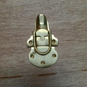 May include: A gold-toned metal latch with a rounded top and a rectangular opening. The latch has four small holes on the bottom and two on the sides.