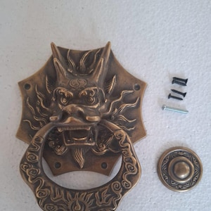 Op de afbeelding: Een messing deurklopper in de vorm van een drakenkop met een ringvormige handgreep. De deurklopper heeft ingewikkelde details en een yin yang symbool op het voorhoofd. Hij wordt geleverd met een ronde decoratieve plaat en schroeven voor installatie.