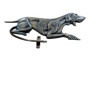May include: A bronze-coloured metal dog sculpture in a reclining position, with a curved tail and a mounting bracket. The dog has detailed features and is likely a wall-mounted decorative item.