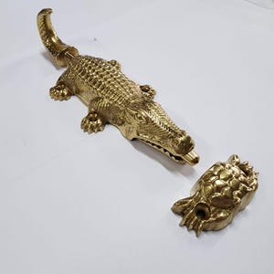 Puede incluir: Un tope de puerta con forma de cocodrilo de color dorado con una superficie texturizada. El tope de puerta está en dos piezas, el cocodrilo y una pieza separada que encaja en la boca del cocodrilo.