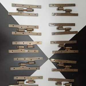 Puede incluir: Colección de bisagras antiguas de color latón. Cada bisagra tiene una base rectangular con múltiples orificios para tornillos y un cilindro giratorio. Las bisagras están dispuestas sobre una superficie a cuadros en blanco y negro.