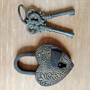 May include: A heart-shaped padlock with an ornate design and a keyhole. The padlock is made of brass and has a dark patina. It comes with two keys with intricate designs.