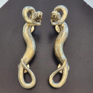 May include: A pair of gold-toned snake door handles with detailed scales and open mouths. The handles are designed to be mounted on a door.