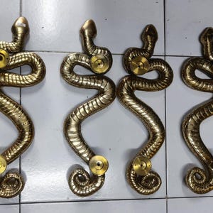 May include: Six gold-coloured snake-shaped door handles. Each handle features a coiled body, a raised head, and a circular brass-coloured knob. The handles are arranged in two rows of three.