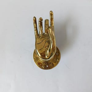 May include: A golden-colored, hand-shaped decorative object, with the index and pinky fingers extended. The hand is mounted on a circular base with an ornate design. The object is likely made of brass or a similar metal.