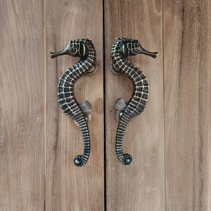 May include: A pair of ornate door handles shaped like seahorses. The handles are made of brass and have a dark finish. They are mounted on a wooden door.