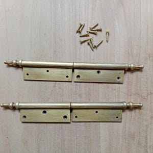 May include: Two brass hinges with decorative finials and a set of screws. The hinges are designed for heavy doors and have a traditional style.