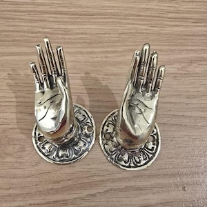 Könnte beinhalten: Zwei türgriffe in Form von Händen in Messingfarbe. Jede Hand ist in einer Geste des Angebots, montiert auf einer dekorativen kreisförmigen Basis. Das Metall hat eine polierte Oberfläche, die das Licht reflektiert. Der Hintergrund ist eine hellbraune Holzoberfläche.