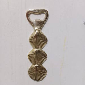 May include: A silver bottle opener with a seashell design. The opener has a loop at the top and three stacked seashell shapes along the handle. The opener is made of metal and has a shiny finish. The design is reminiscent of the coast.