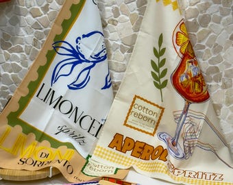 Puede incluir: Paños de cocina triangulares con diseños de temática italiana. Uno presenta un diseño floral azul con texto "Limoncello Spritz", y el otro muestra una ilustración de Aperol Spritz. También se ve una pila de toallas más pequeñas con varios patrones.