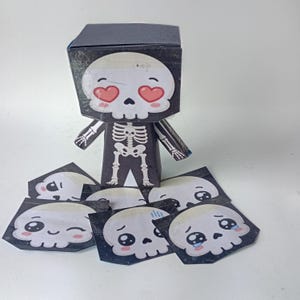 Peut inclure: Une figurine squelette en papier avec une grande tête carrée représentant un crâne blanc aux yeux en forme de cœur. Le corps est noir avec une impression de cage thoracique blanche. Plusieurs visages de crânes en papier sont disposés en dessous.