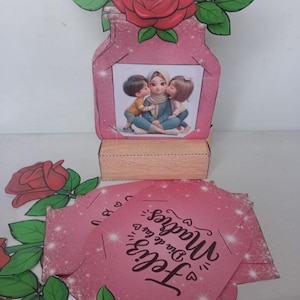 Pode incluir: Um vaso de papel rosa com rosas de papel vermelho e folhas verdes. O vaso tem uma imagem de uma mãe com duas crianças. Cartões rosa com o texto "Feliz Dia de las Madres" estão na frente.
