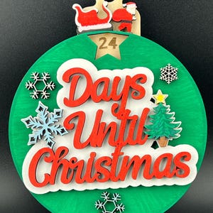 May include: A green circular wooden ornament with the text "Days Until Christmas" in red lettering. The ornament has a star with the number 24 in it, a small Christmas tree, and snowflakes.