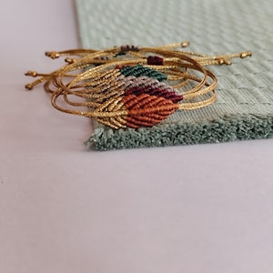 May include: Several gold-coloured macrame bracelets with leaf-shaped details in various colours. The bracelets have woven patterns in red, orange, green, and grey. They are arranged on a light green textured surface.