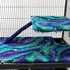 May include: A black metal cage with two levels, both covered in a blue, green, and purple fabric with a white star pattern. The top level has a ramp leading up to it.