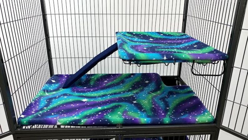 May include: A black metal cage with two levels, both covered in a blue, green, and purple fabric with a white star pattern. The top level has a ramp leading up to it.