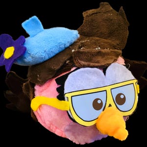 May include: A pink plush bird with brown hair, blue sunglasses, and a blue beret with a purple flower. The bird has a yellow beak and a light blue eye.