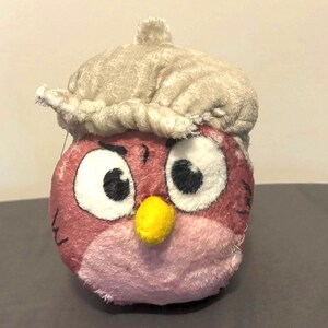 May include: A plush toy shaped like an owl with a light brown hat. The owl has large white eyes with black pupils, a yellow beak, and pink and red feathers. The toy is made of soft, fuzzy material.