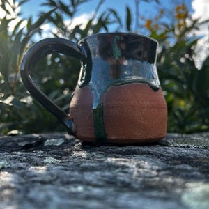 May include: A small, handmade ceramic mug with a brown body and a dark green and black glaze. The mug has a rounded shape and a handle.