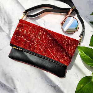 May include: A red velvet clutch with a black leather flap and a gold zipper. The clutch has a long black strap and is sitting on a white marble surface.