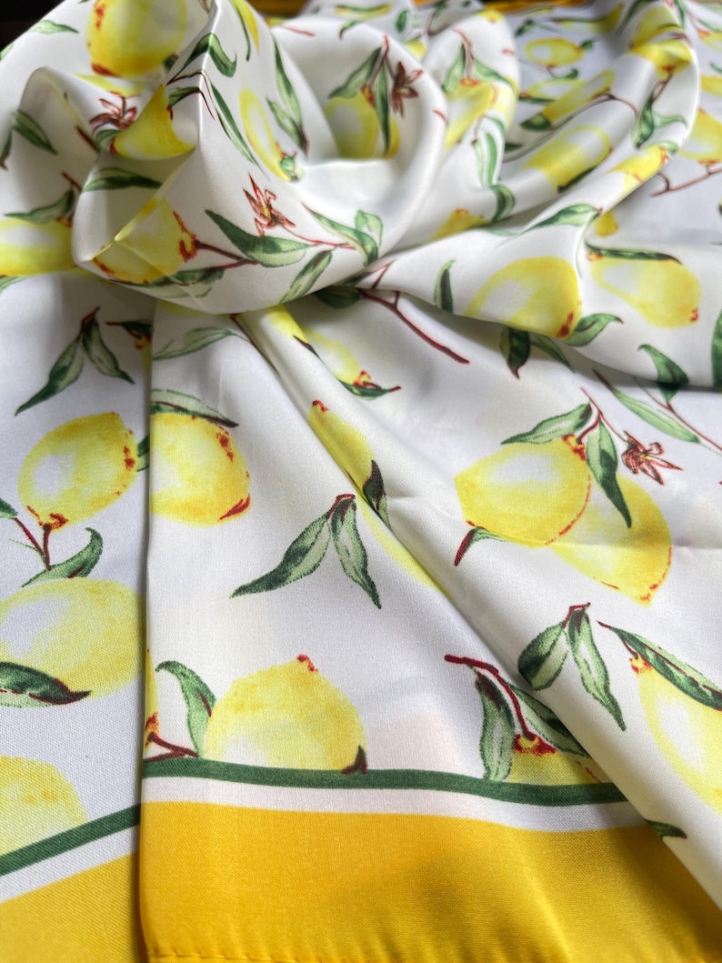 May include: White silk scarf with a yellow lemon print and a yellow border. The lemons are arranged in a repeating pattern on the scarf.