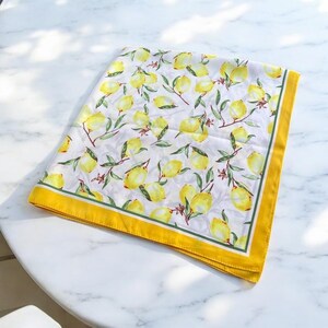May include: A white square scarf with a yellow border and a lemon print. The lemons are yellow with green leaves and stems.