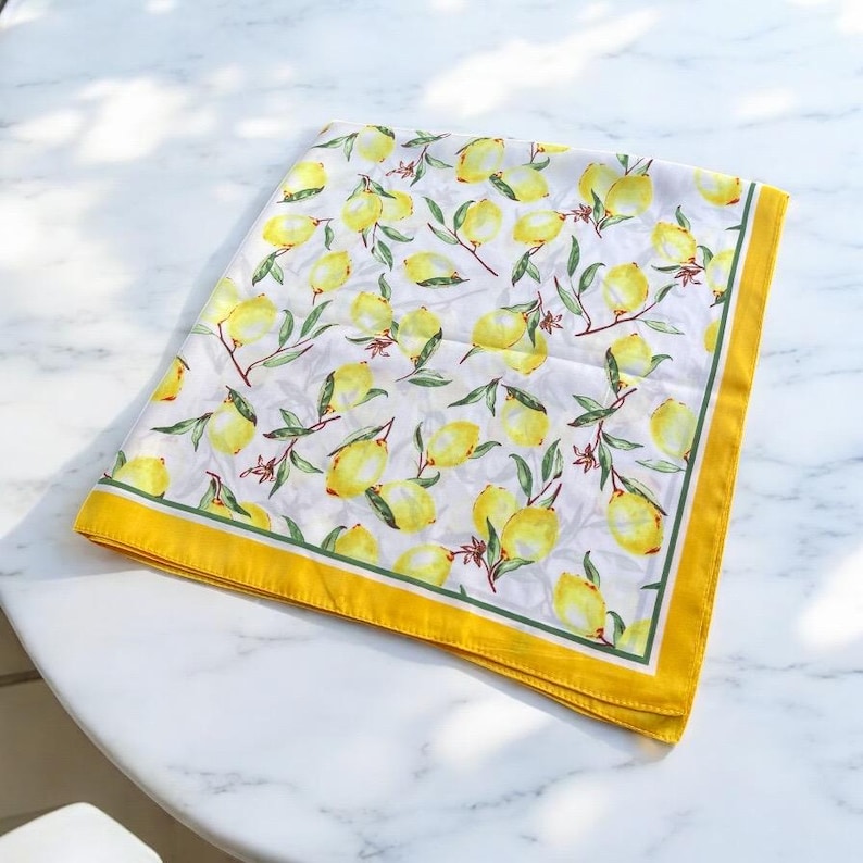May include: A white square scarf with a yellow border and a lemon print. The lemons are yellow with green leaves and stems.