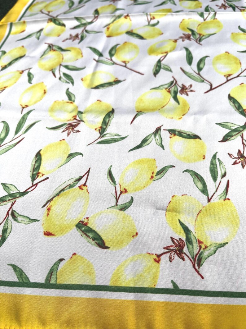 May include: White fabric with a yellow border and a repeating pattern of yellow lemons with green leaves.
