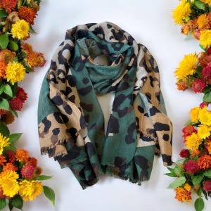 May include: A green and brown leopard print scarf with a fringed edge. The scarf is folded in half and laid flat on a white surface.